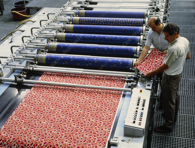 extile printing. Workers assessing the colours on a textile sheet after printing. The textile pattern has been printed using the rollers of a rotary printer (at upper centre). Each of the rollers print a single colour onto the textile, each forming a part of the completed pattern. Photographed in Germany.
